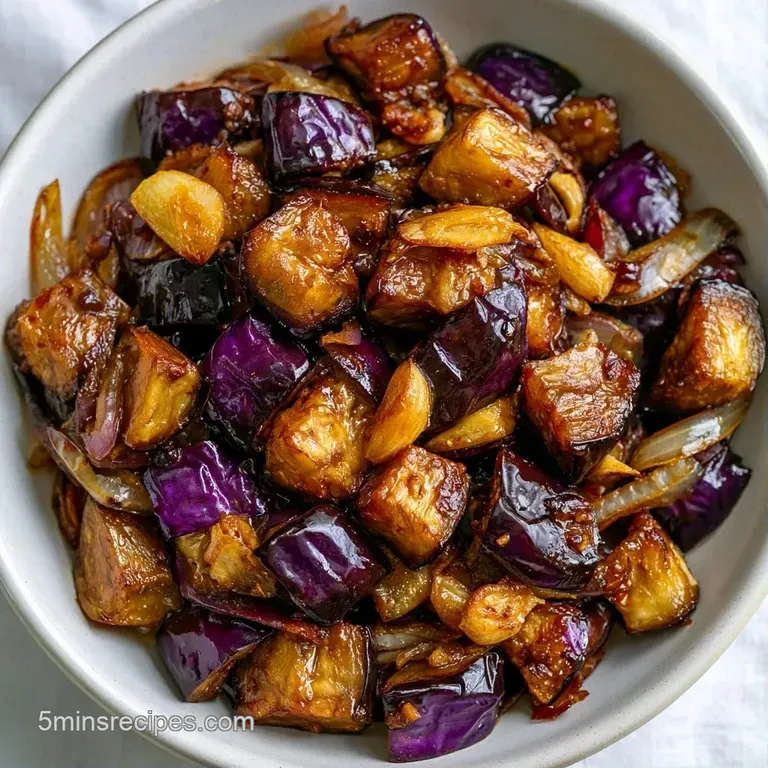 A tidy pile of tender, browned eggplant chunks on a white platter, topped with toasted garlic and bright greenery.